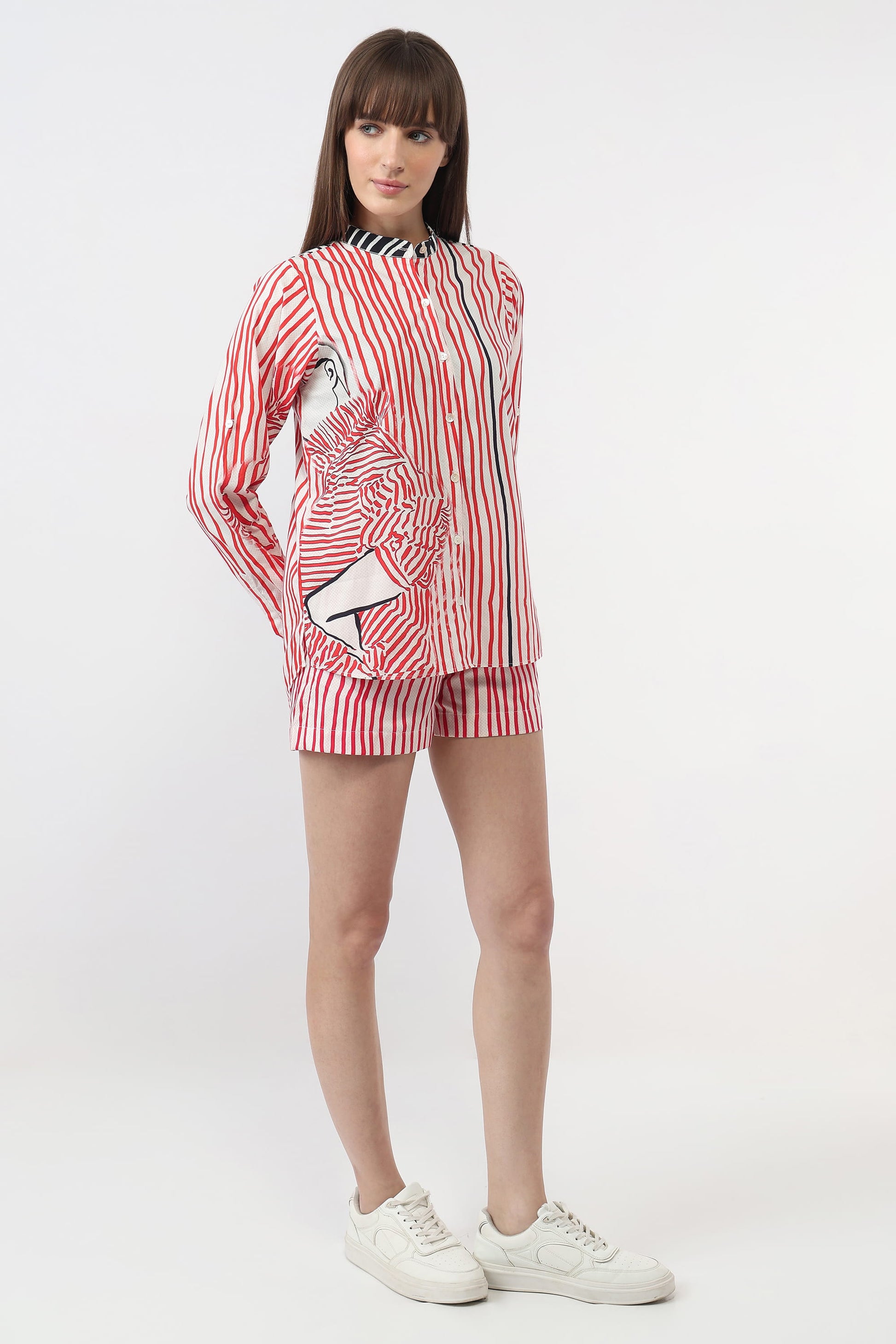 A model wearing a matching red and white striped lounge set consisting of a long-sleeve button-up shirt and shorts, paired with white sneakers, against a plain white studio background.