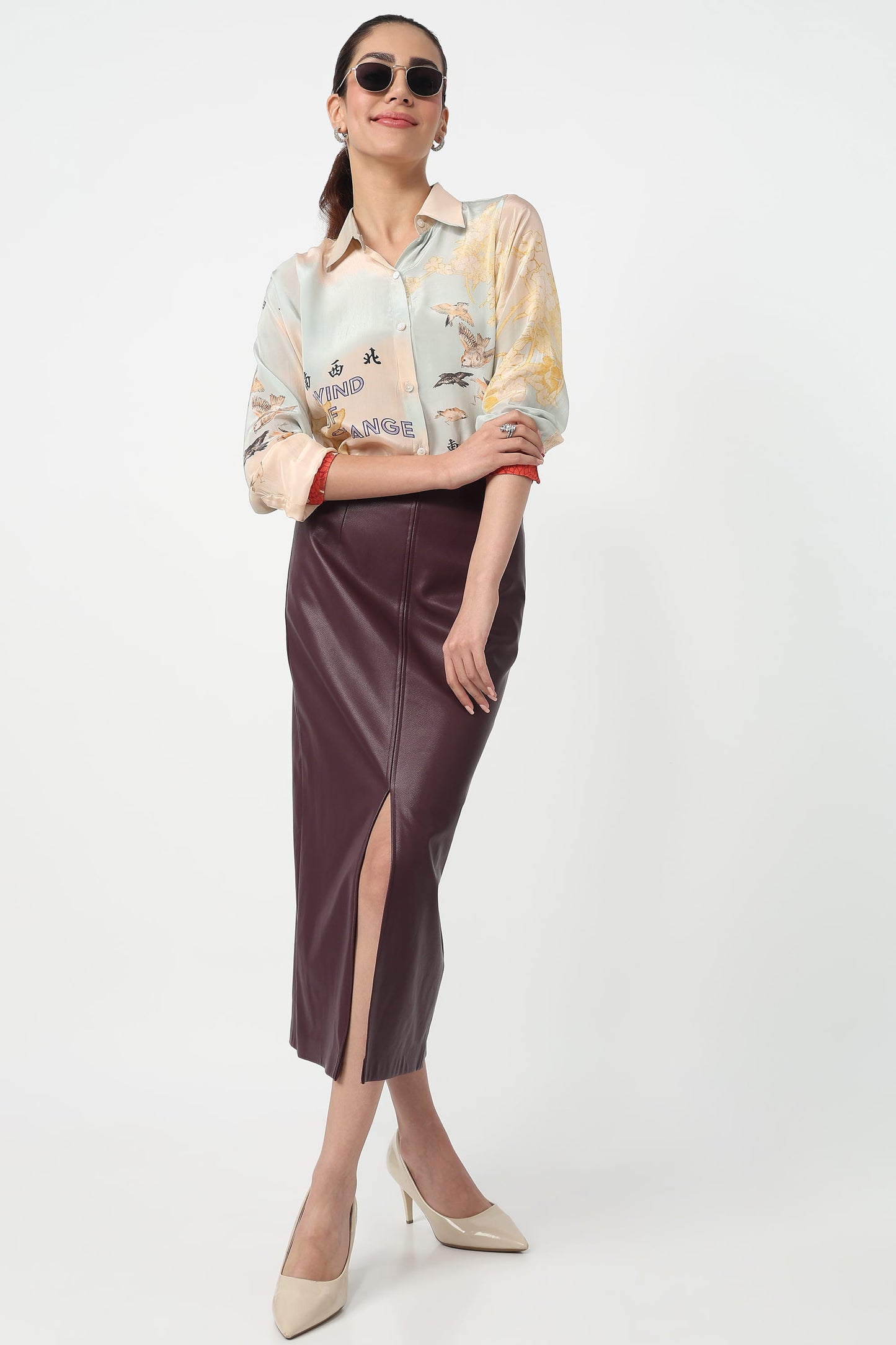 A woman modeling a long-sleeve collared shirt with a multicolor abstract print and a dark brown faux-leather midi skirt with a front slit, paired with beige heels against a light gray studio background.