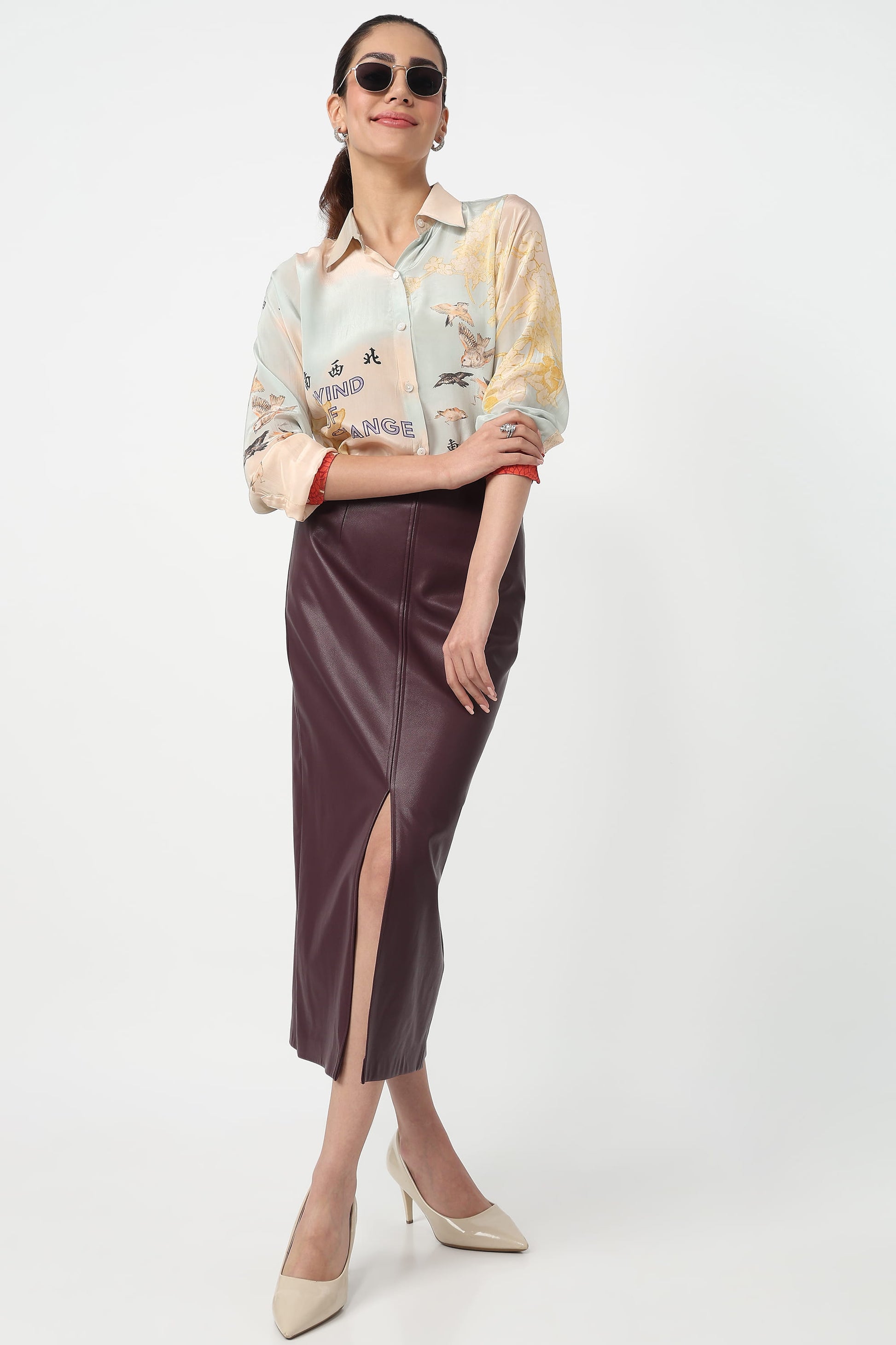 A woman modeling a long-sleeve collared shirt with a multicolor abstract print and a dark brown faux-leather midi skirt with a front slit, paired with beige heels against a light gray studio background.