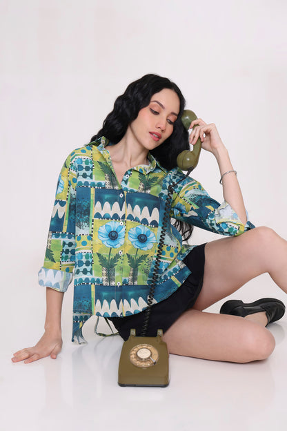 A woman sits on a white floor wearing a colorful patterned button-up shirt with blue, green, and yellow floral and geometric designs, paired with black shorts. She holds a vintage olive green rotary telephone to her ear while looking down at it.