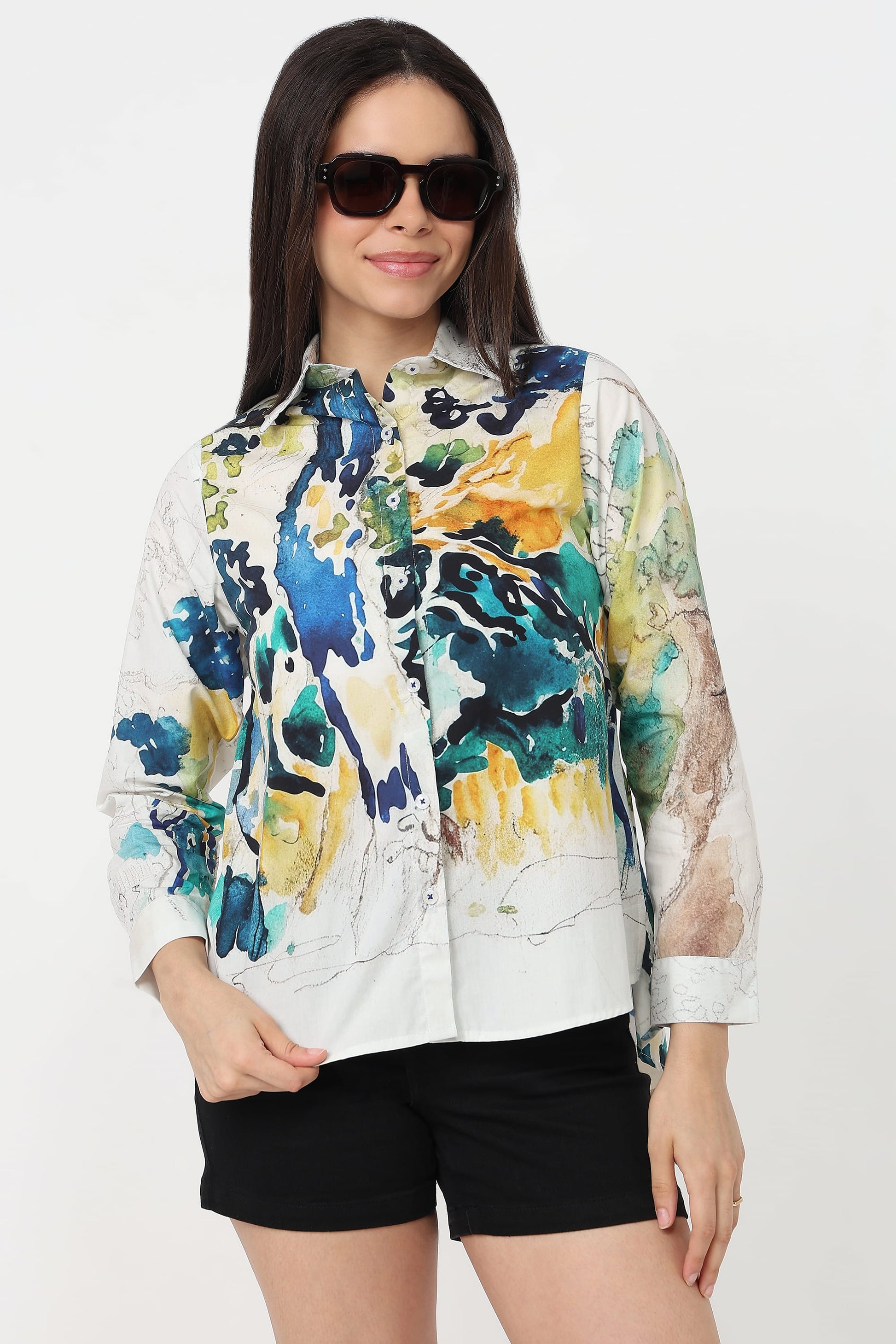 A woman wearing a white button-up shirt with a vibrant abstract watercolour print in blue, green, yellow, and black, paired with black shorts against a plain white background.