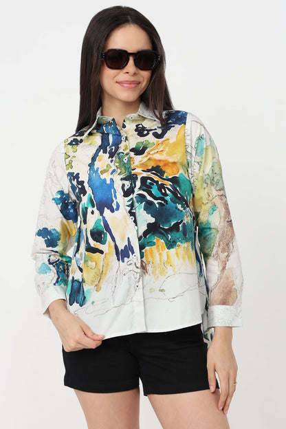 A woman wearing a white button-up shirt with a vibrant abstract watercolour print in blue, green, yellow, and black, paired with black shorts against a plain white background.