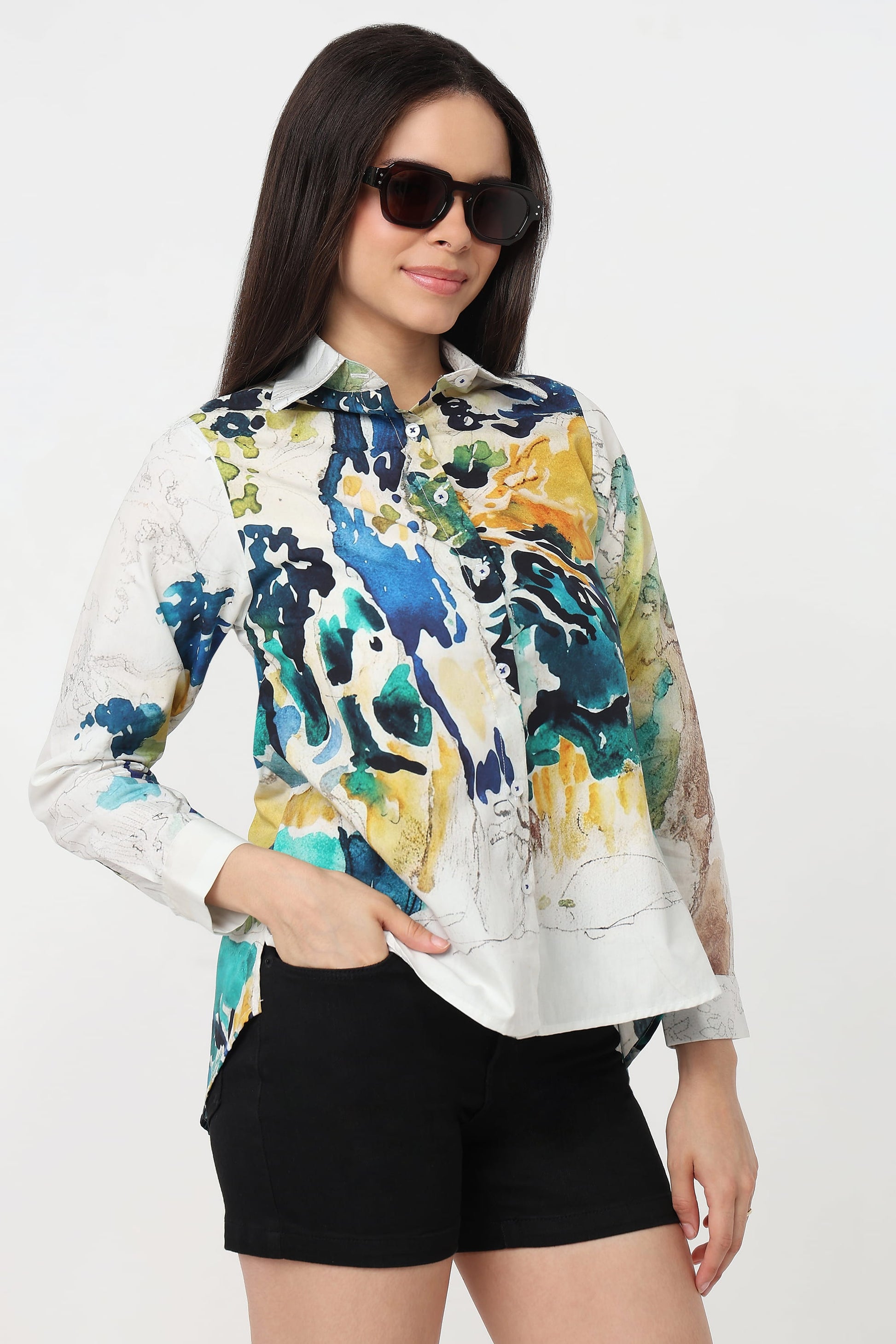 A woman wearing a long-sleeve button-up shirt with a vibrant abstract watercolour print in blue, green, yellow, and white, paired with black shorts against a plain white background.