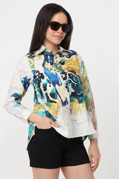 A woman wearing a long-sleeve button-up shirt with a vibrant abstract watercolour print in blue, green, yellow, and white, paired with black shorts against a plain white background.