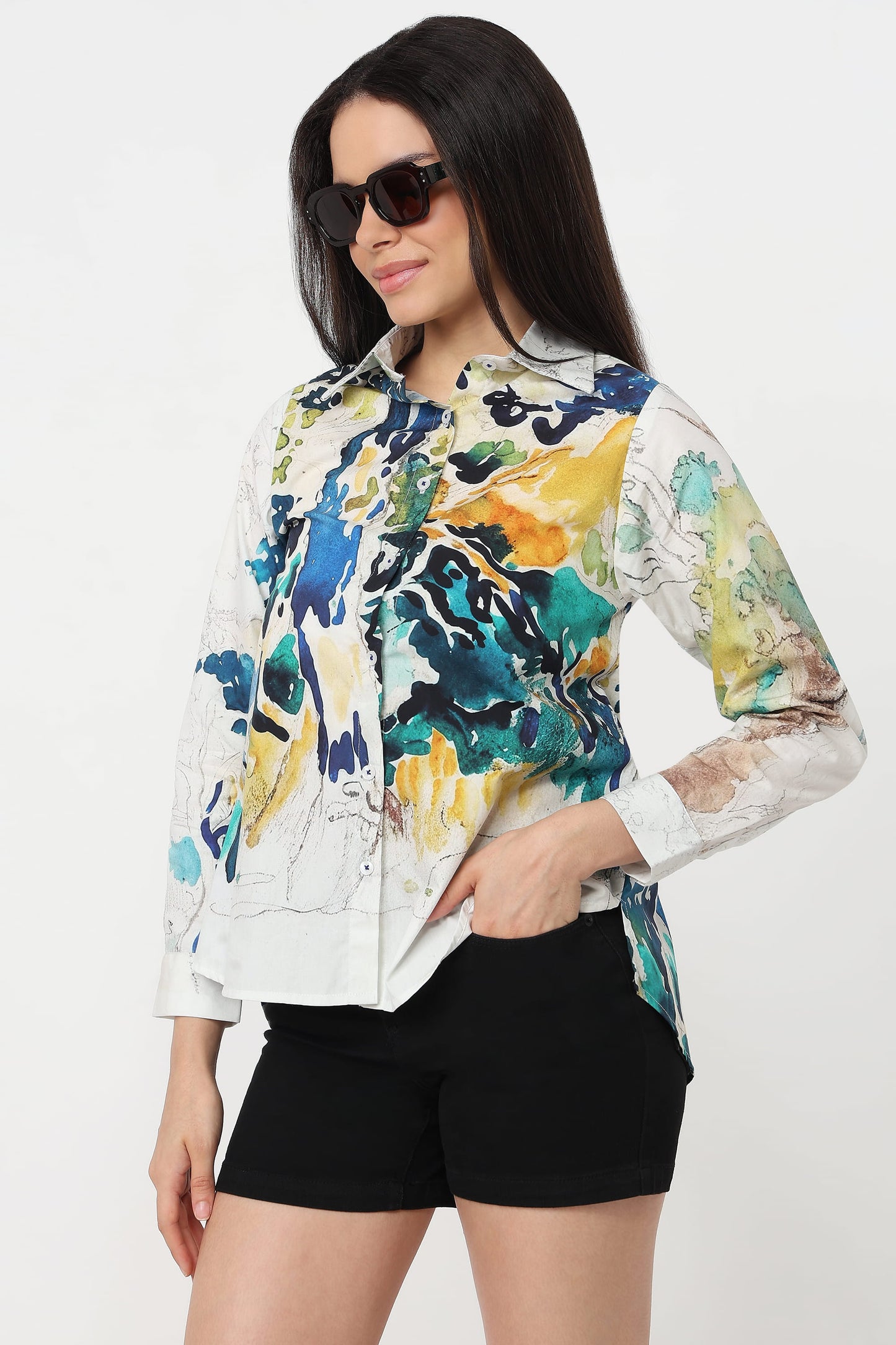 A woman wearing a long-sleeve button-up shirt with an abstract watercolour print in blue, yellow, green, and white, paired with black shorts against a plain white background.