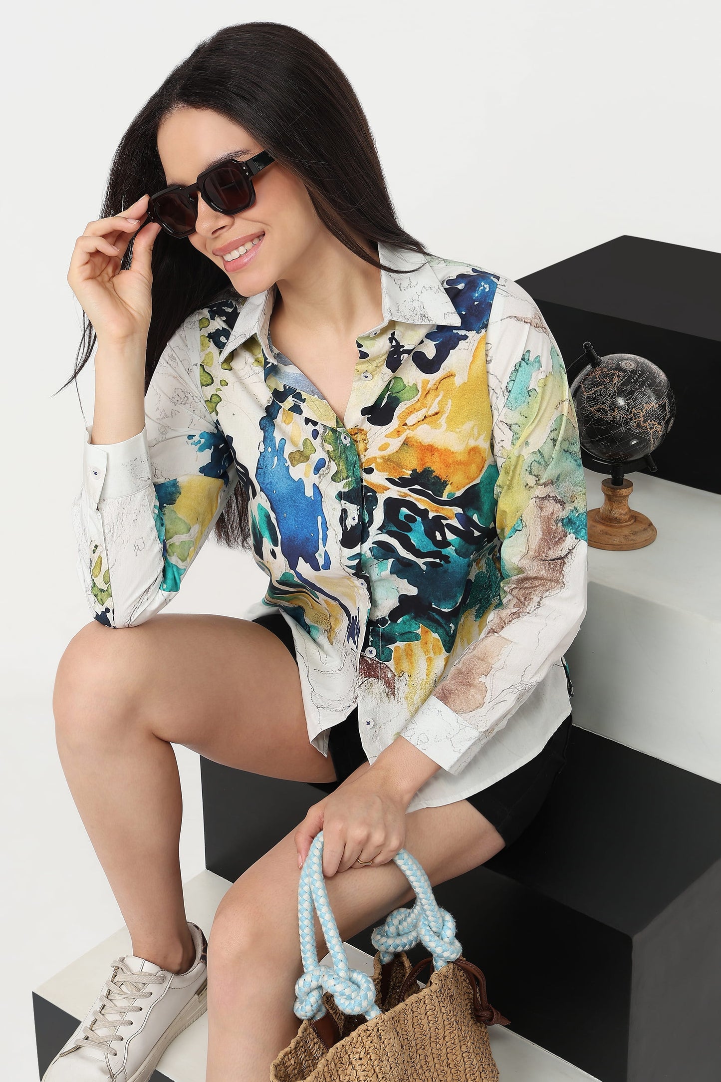 A woman wearing a white button-up shirt with a vibrant blue, yellow, and green abstract watercolor print, styled with black shorts, white sneakers, and sunglasses while sitting on a black and white geometric block.