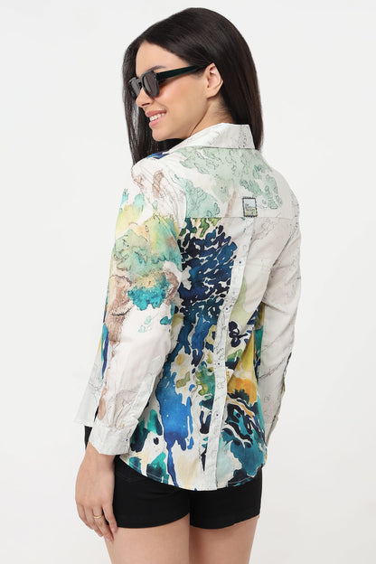 A woman wearing a long-sleeve button-up shirt with a colorful abstract watercolor print in blue, green, yellow, and beige on an off-white base, paired with black shorts and sunglasses, shown from a three-quarters back view against a plain white background.