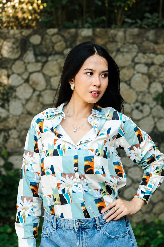 A woman wearing a long-sleeve button-up shirt with a colorful geometric and floral print, paired with blue jeans, standing outdoors in front of a stone wall.