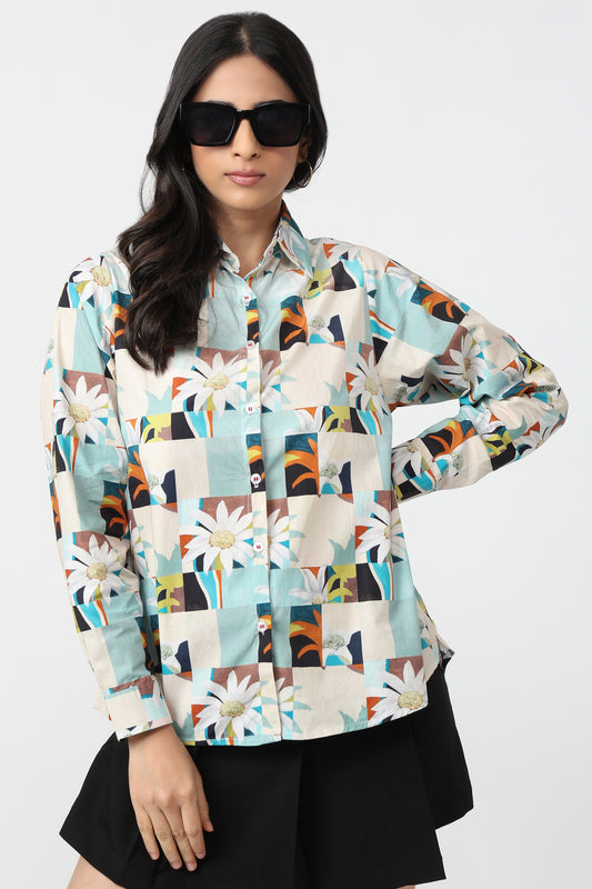 A woman modeling a long-sleeve button-up shirt with a colorful geometric and floral patchwork print, paired with black shorts and black sunglasses, against a plain light gray background.