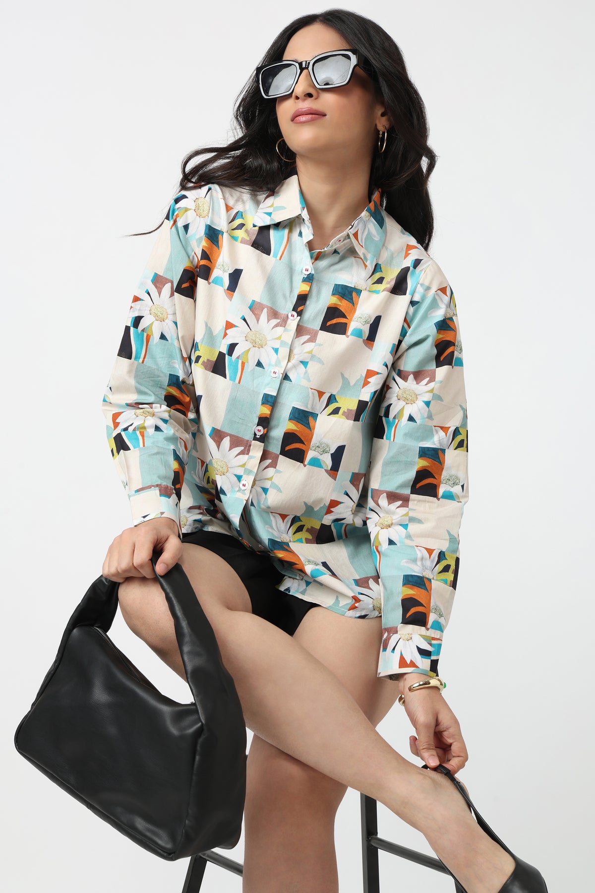 A woman wearing a long-sleeve button-up shirt with a colorful geometric and floral patchwork print, styled with black shorts, black sunglasses, and a black shoulder bag, sitting on a stool against a white studio background.