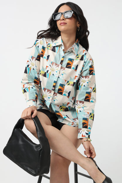 A woman wearing a long-sleeve button-up shirt with a colorful geometric and floral patchwork print, styled with black shorts, black sunglasses, and a black shoulder bag, sitting on a stool against a white studio background.