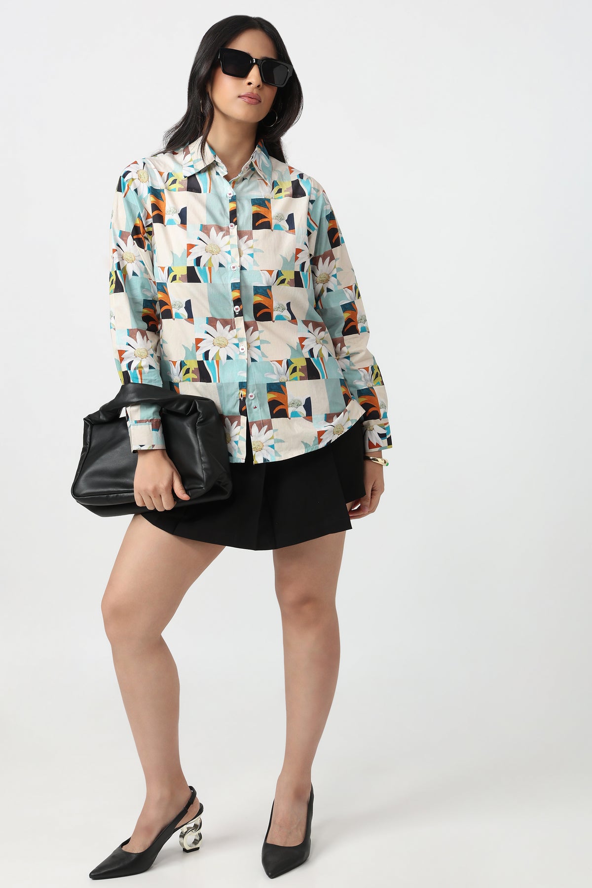 A woman modeling a long-sleeve button-up shirt with a colorful geometric and floral patchwork print, paired with a black mini skirt, black handbag, and black pointed-toe heels against a light gray studio background.