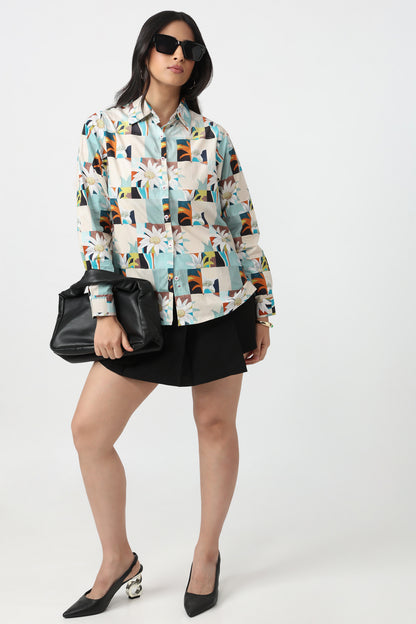 A woman modeling a long-sleeve button-up shirt with a colorful geometric and floral patchwork print, paired with a black mini skirt, black handbag, and black pointed-toe heels against a light gray studio background.