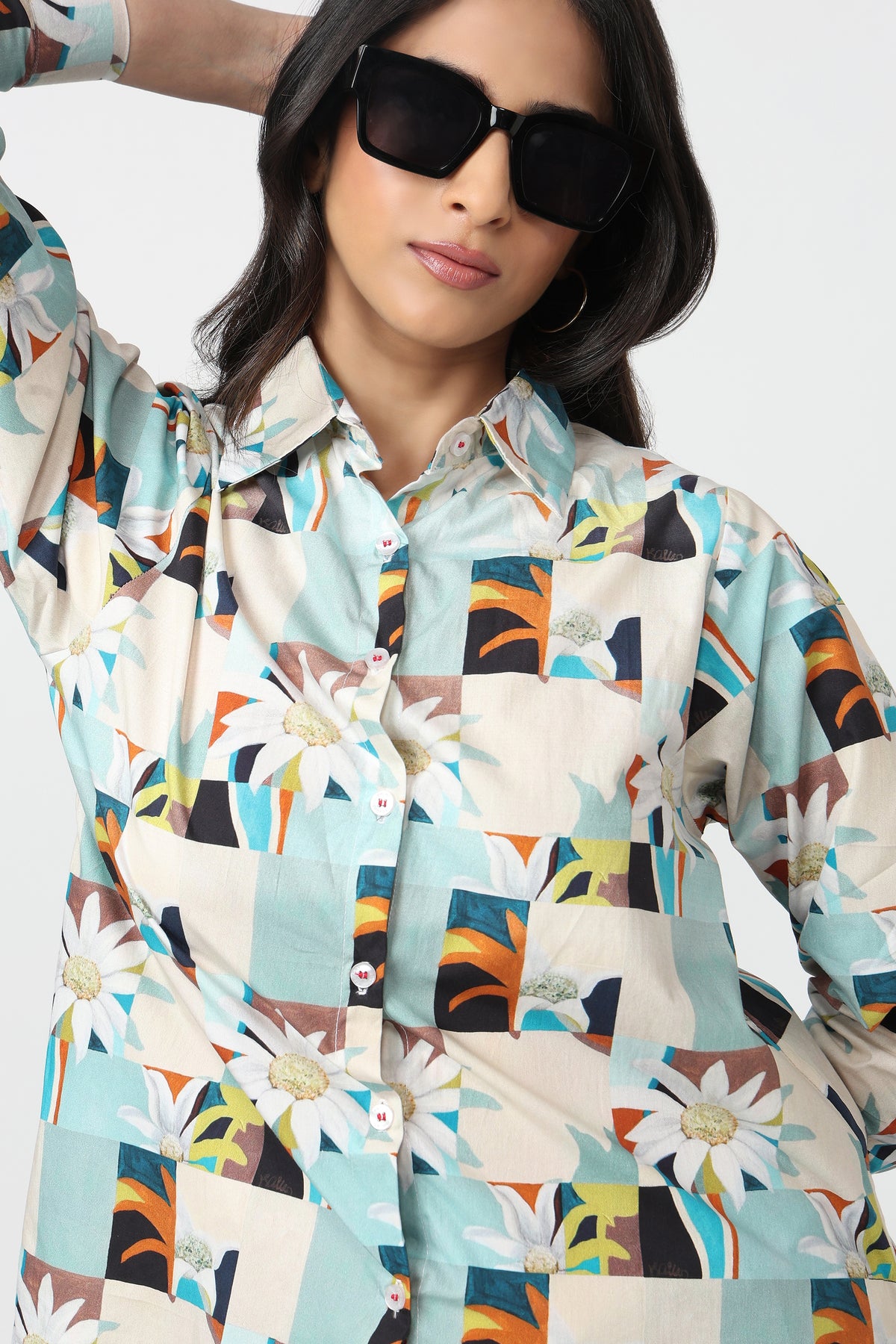 A woman wearing a long-sleeve button-up shirt with a colorful geometric patchwork print featuring daisies, abstract shapes, and tropical motifs in light blue, beige, orange, brown, and white. She is also wearing black sunglasses against a plain light gray background.