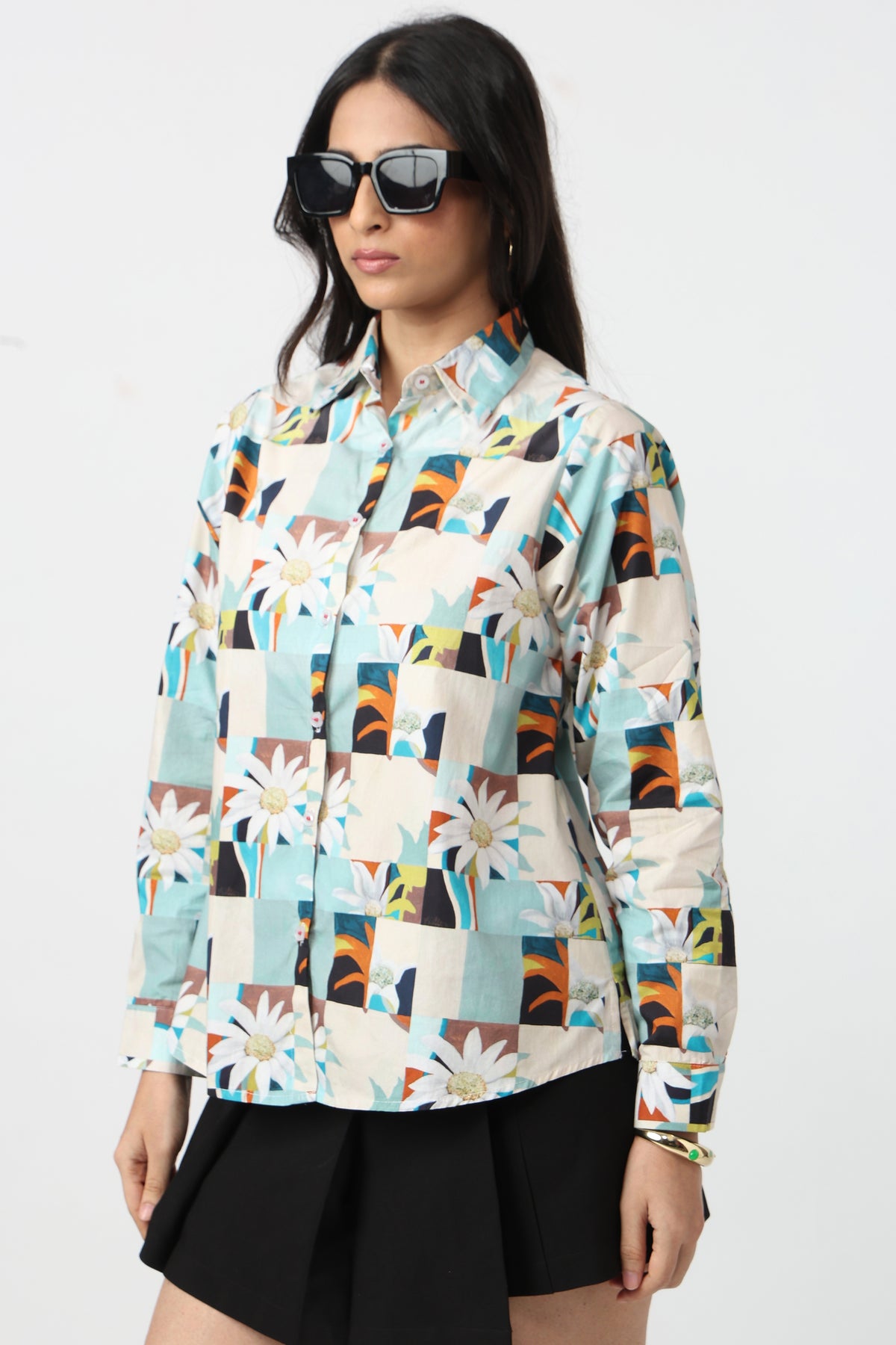 A woman modeling a long-sleeve button-up shirt with a colorful patchwork print featuring daisies, geometric shapes, and abstract blocks in light blue, beige, orange, brown, and black, paired with black shorts and sunglasses against a plain white background.
