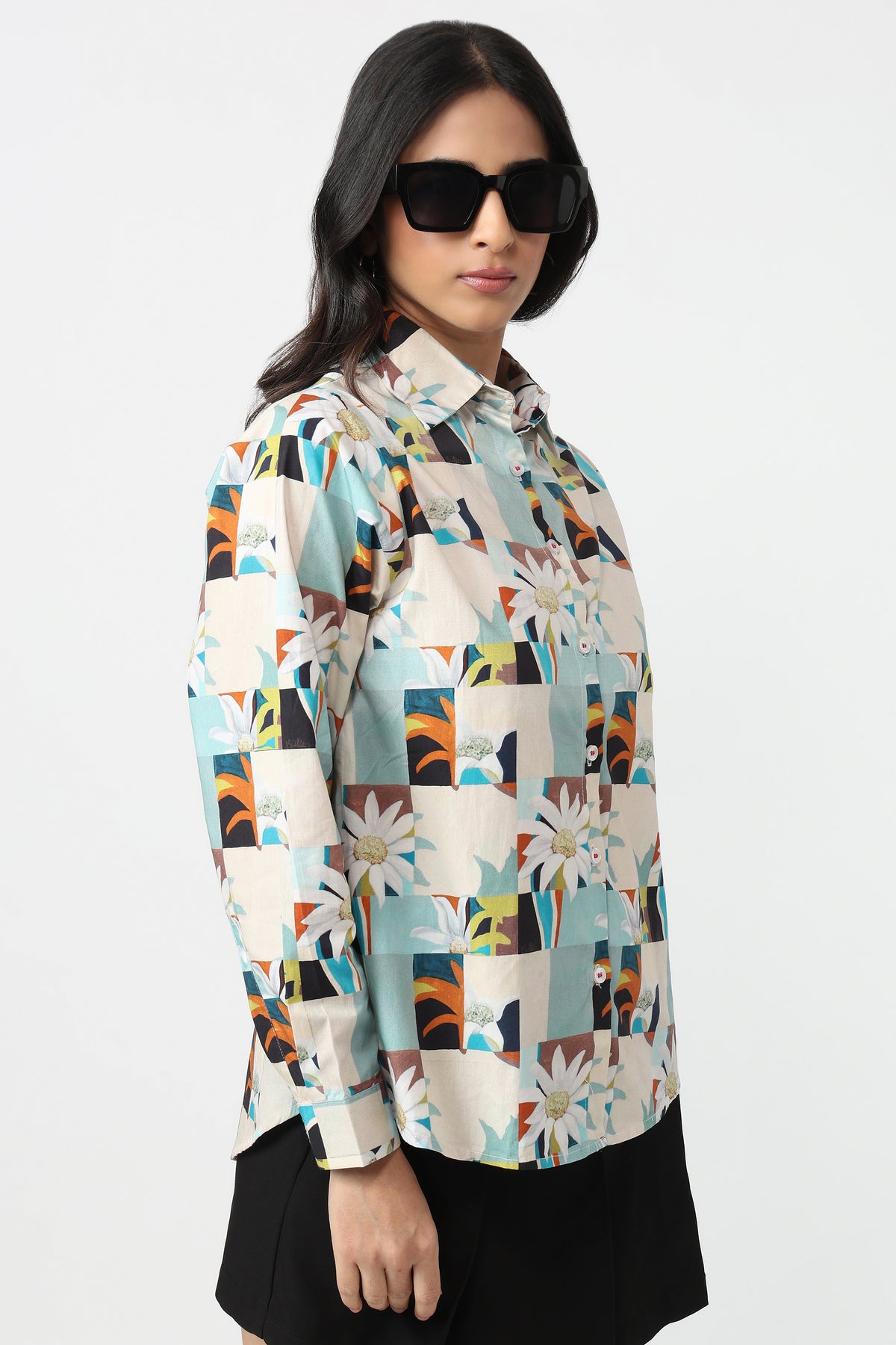 A woman modeling a long-sleeve button-up shirt with a colorful patchwork floral print, paired with black sunglasses and a black skirt, against a plain white studio background.