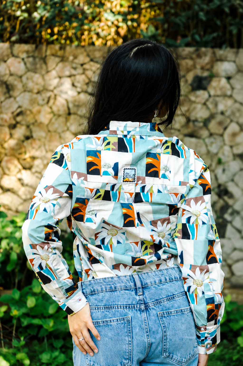 A woman viewed from behind wearing a long-sleeve button-up shirt with a colorful geometric and floral patchwork print, paired with light blue denim jeans, set against a stone wall background.