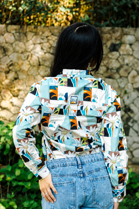 A woman viewed from behind wearing a long-sleeve button-up shirt with a colorful geometric and floral patchwork print, paired with light blue denim jeans, set against a stone wall background.