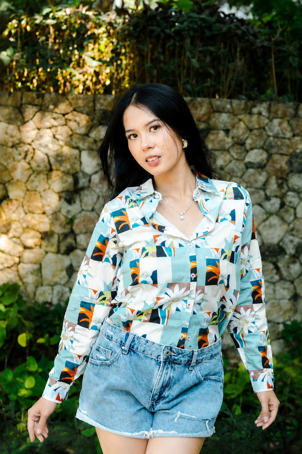 A woman wearing a long-sleeve button-up shirt with a colorful geometric patchwork pattern in shades of blue, orange, black, and white, paired with light-wash denim shorts, standing outdoors in front of a stone wall and green foliage.