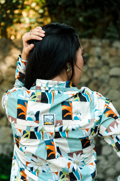 A woman viewed from behind wearing a hooded jacket with a colorful geometric and floral print in light blue, white, orange, black, and green. She is outdoors with a blurred stone wall and foliage in the background.