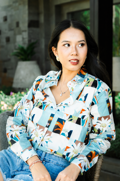 A woman wearing a long-sleeve button-up shirt with a colorful geometric and floral patchwork print, paired with blue jeans, sitting in an outdoor patio setting.