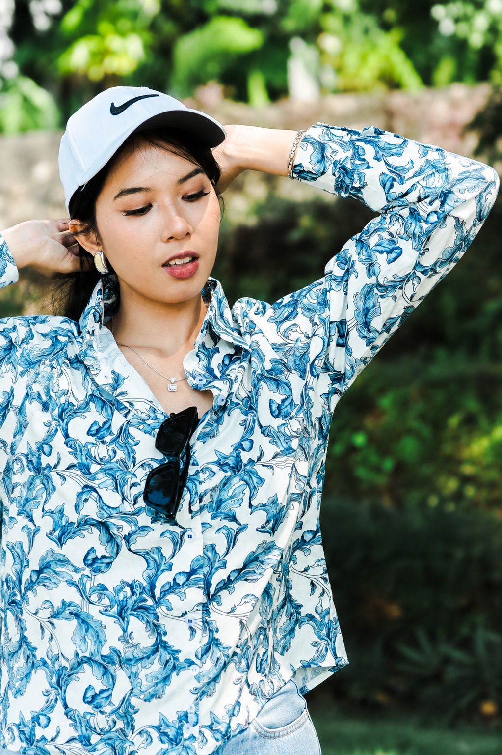 A woman wearing a white long-sleeve button-up shirt with a blue abstract swirl pattern, a white Nike cap, and sunglasses hanging from the collar, posing outdoors against a blurred green background.