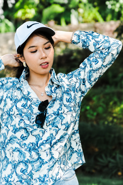 A woman wearing a white long-sleeve button-up shirt with a blue abstract swirl pattern, a white Nike cap, and sunglasses hanging from the collar, posing outdoors against a blurred green background.