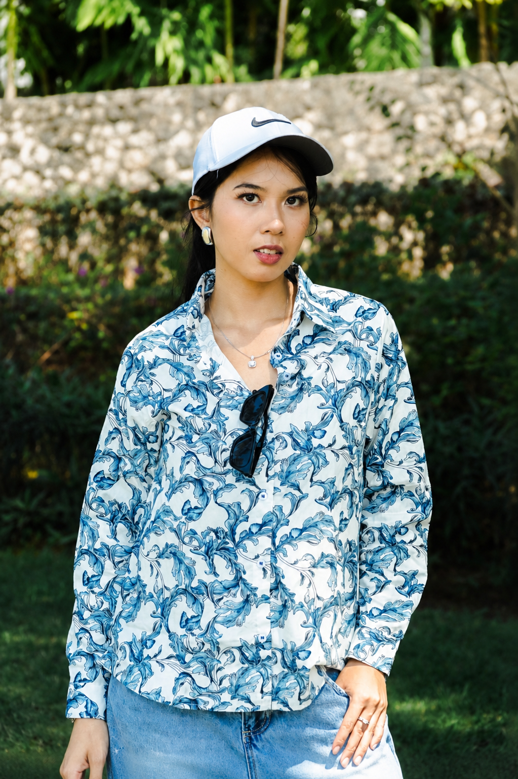 A woman wearing a blue and white abstract patterned button-up shirt, a white Nike cap, and sunglasses hanging from her collar, standing outdoors in front of a stone wall and green foliage.