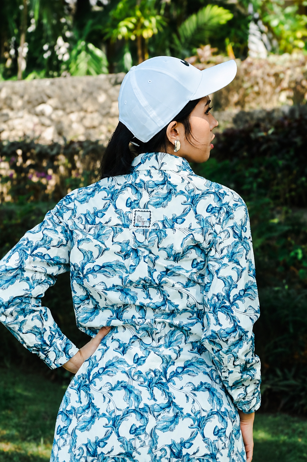 A woman wearing a white baseball cap and a long-sleeve collared shirt with a blue and white abstract swirl pattern, photographed outdoors against a blurred green garden background.