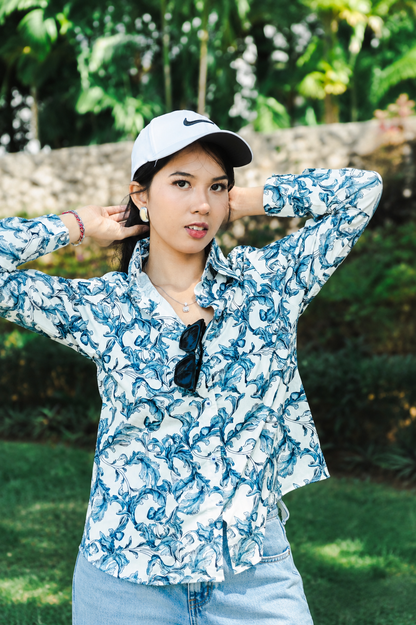 A woman wearing a white and blue patterned long-sleeve button-up shirt, a white Nike cap, and light blue jeans, posing outdoors in front of a green garden background.