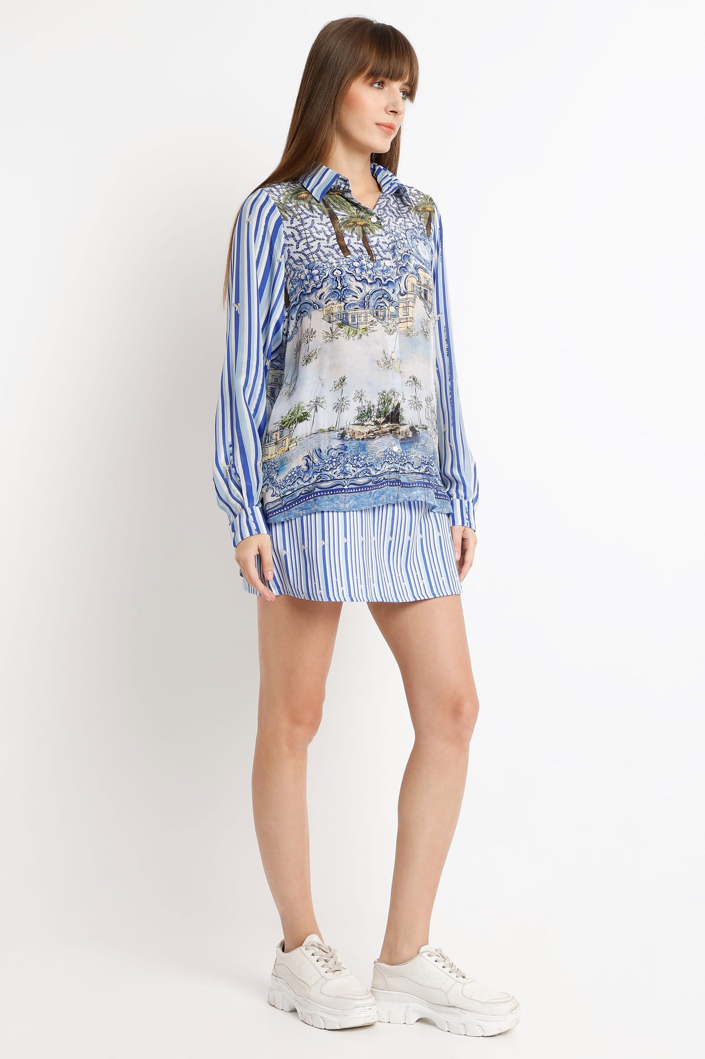 A woman modeling a blue and white striped long-sleeve co-ord set with a scenic landscape print on the front, paired with a matching striped mini skirt and white sneakers, against a plain white studio background.