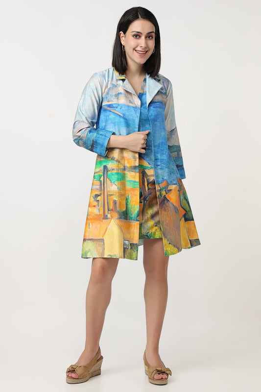 A woman wearing a short-sleeve button-up shirt dress with a colourful abstract art print. The dress features shades of blue, yellow, green, and brown, and is paired with tan wedge sandals against a plain light grey studio background.