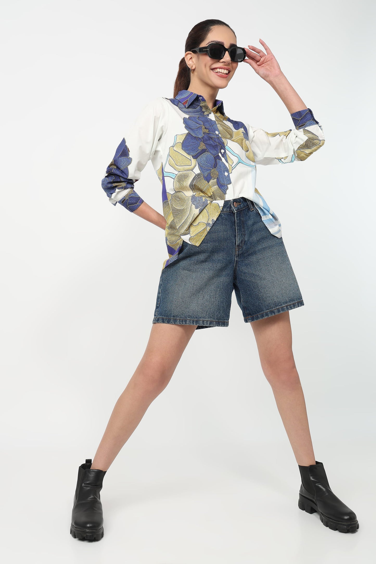 Woman wearing a patterned shirt, denim shorts, and black boots on a white background