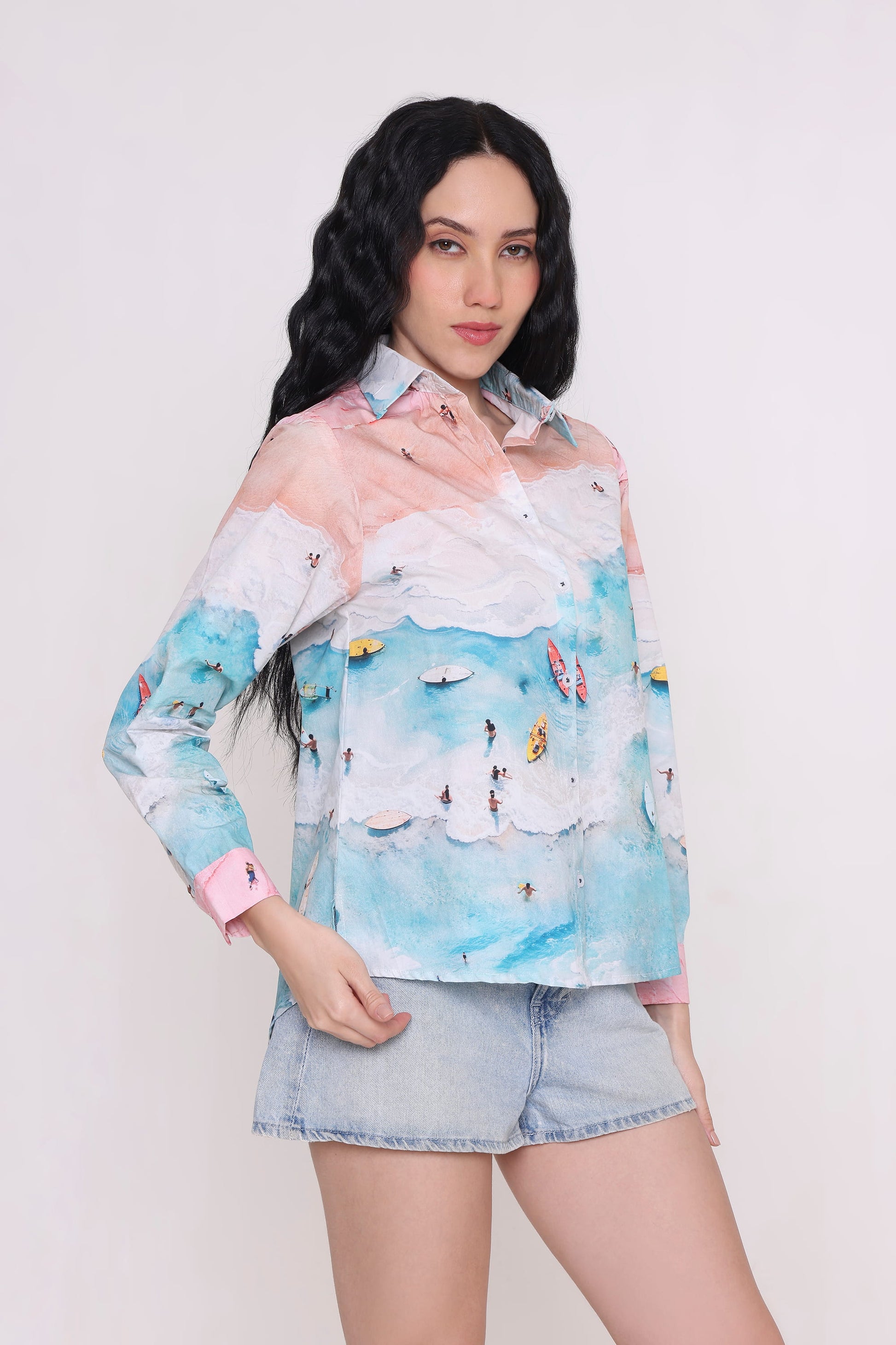 A woman modeling a long-sleeve button-up shirt with a pastel sky and beach print, paired with denim shorts, against a plain light background.