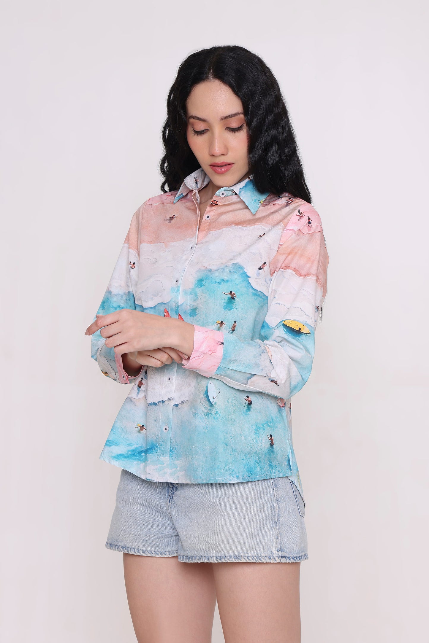 A woman wearing a long-sleeve button-up shirt with a pastel multicolor landscape print featuring pink, blue, and white tones, paired with light denim shorts, against a plain light background.