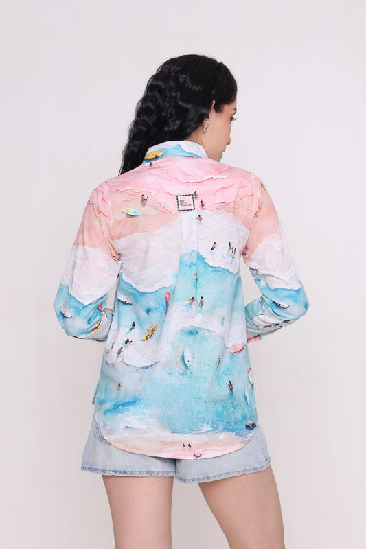 A woman shown from the back wearing a long-sleeve button-up shirt with a pastel beach print in pink, white, and light blue, paired with denim shorts against a plain light background.