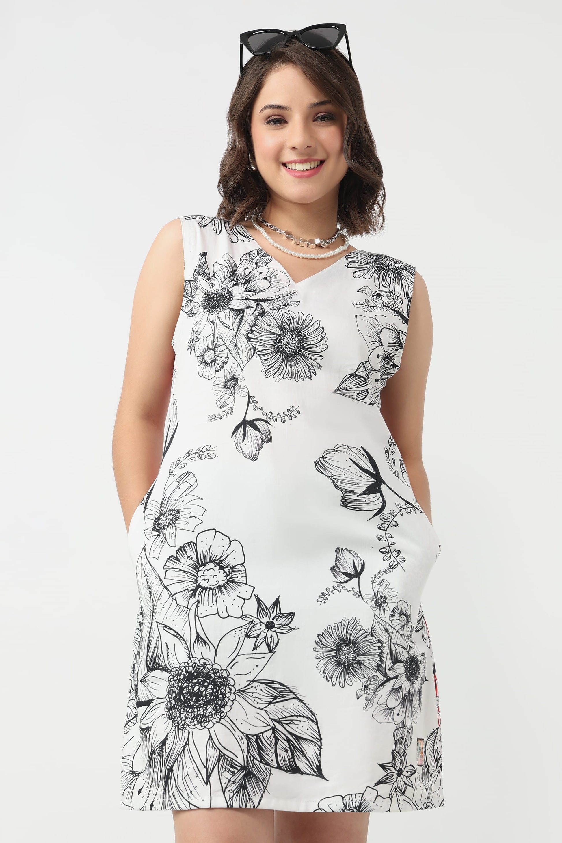 A woman wearing a sleeveless white dress with a black floral line-art print, standing against a light grey studio background. She has sunglasses on her head and layered necklaces.