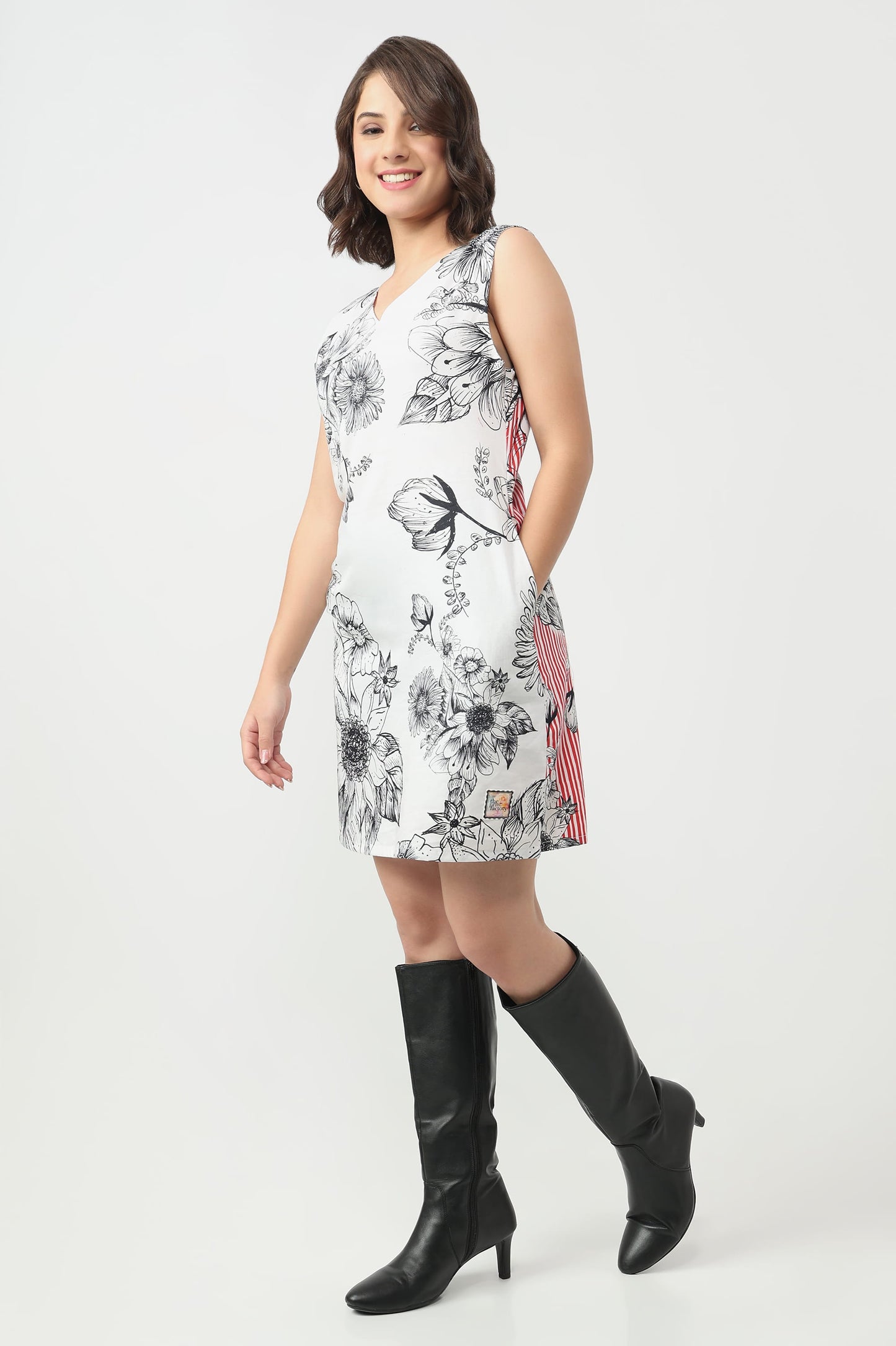 A woman wearing a sleeveless white V-neck dress with a black floral line art print, paired with black knee-high boots, against a light grey studio background.