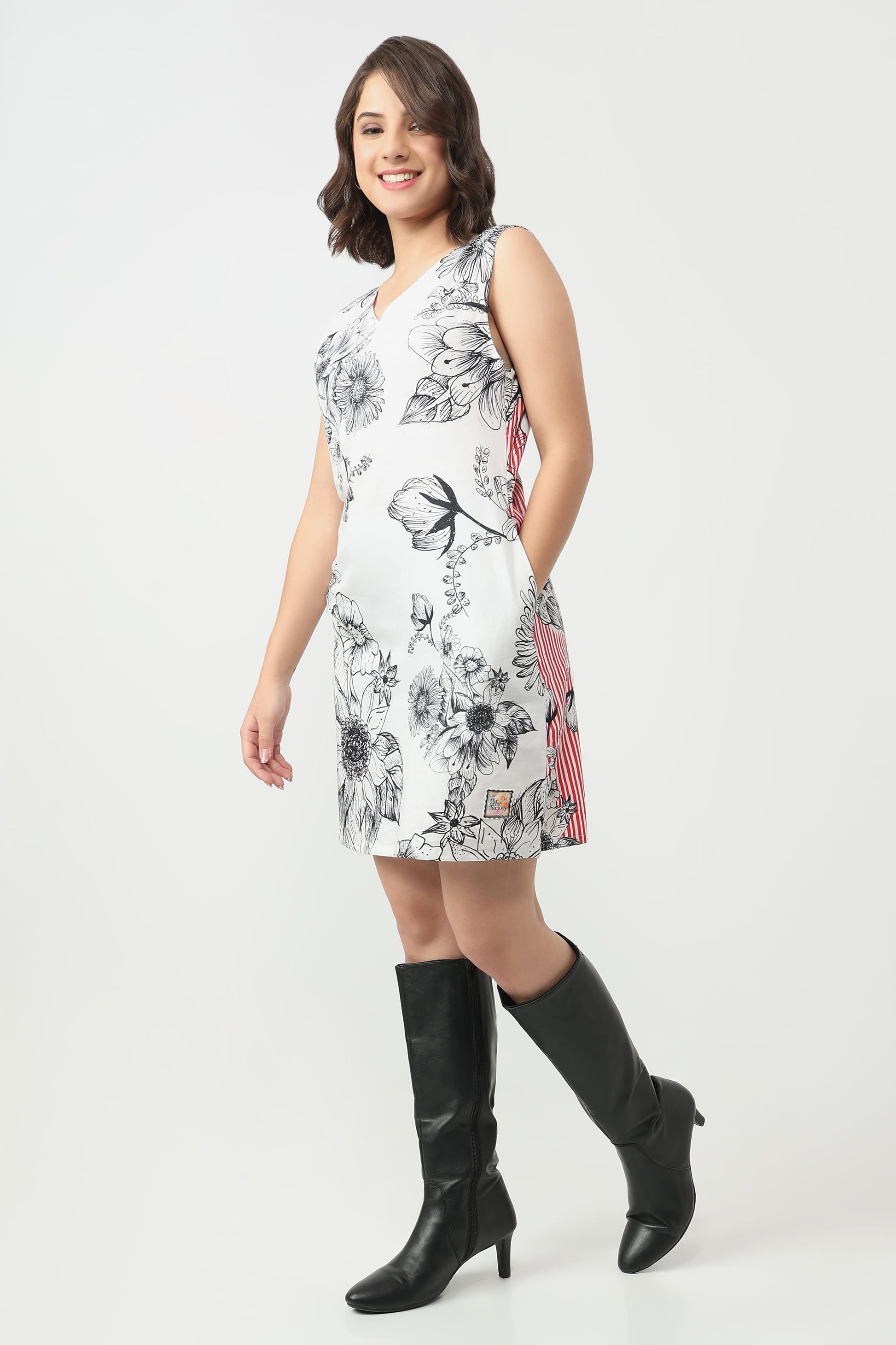 A woman wearing a sleeveless white V-neck dress with a black floral line art print, paired with black knee-high boots, against a light grey studio background.