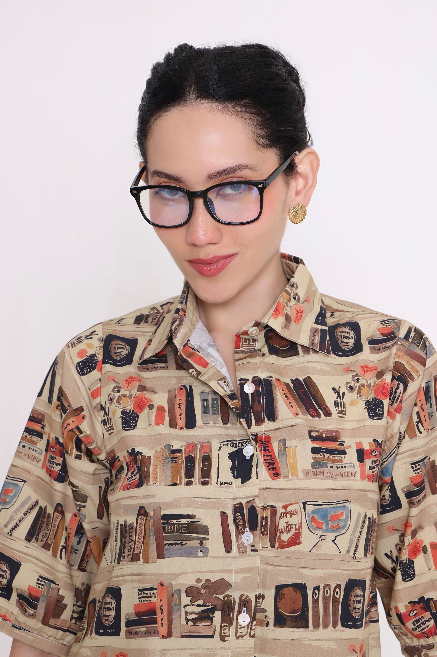 A woman wearing thick black-rimmed glasses and a beige button-up shirt with a detailed graphic print of bookshelves, books, and booksellers, set against a plain white background.