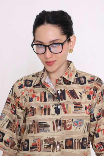 A woman wearing thick black-rimmed glasses and a beige button-up shirt with a detailed graphic print of bookshelves, books, and booksellers, set against a plain white background.