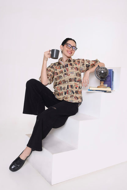 A woman wearing a beige and brown patterned short-sleeve button-up shirt with black wide-leg trousers and black loafers sits on white geometric steps, holding a black mug.