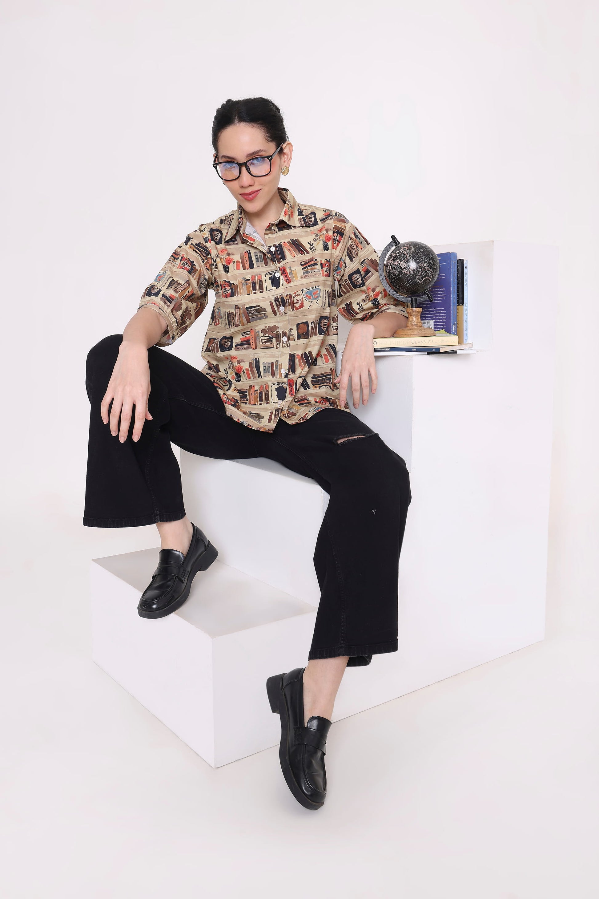 A model wearing a beige and black patterned button-down shirt, black cropped trousers, and black loafers sits on a white geometric platform in a studio setting.
