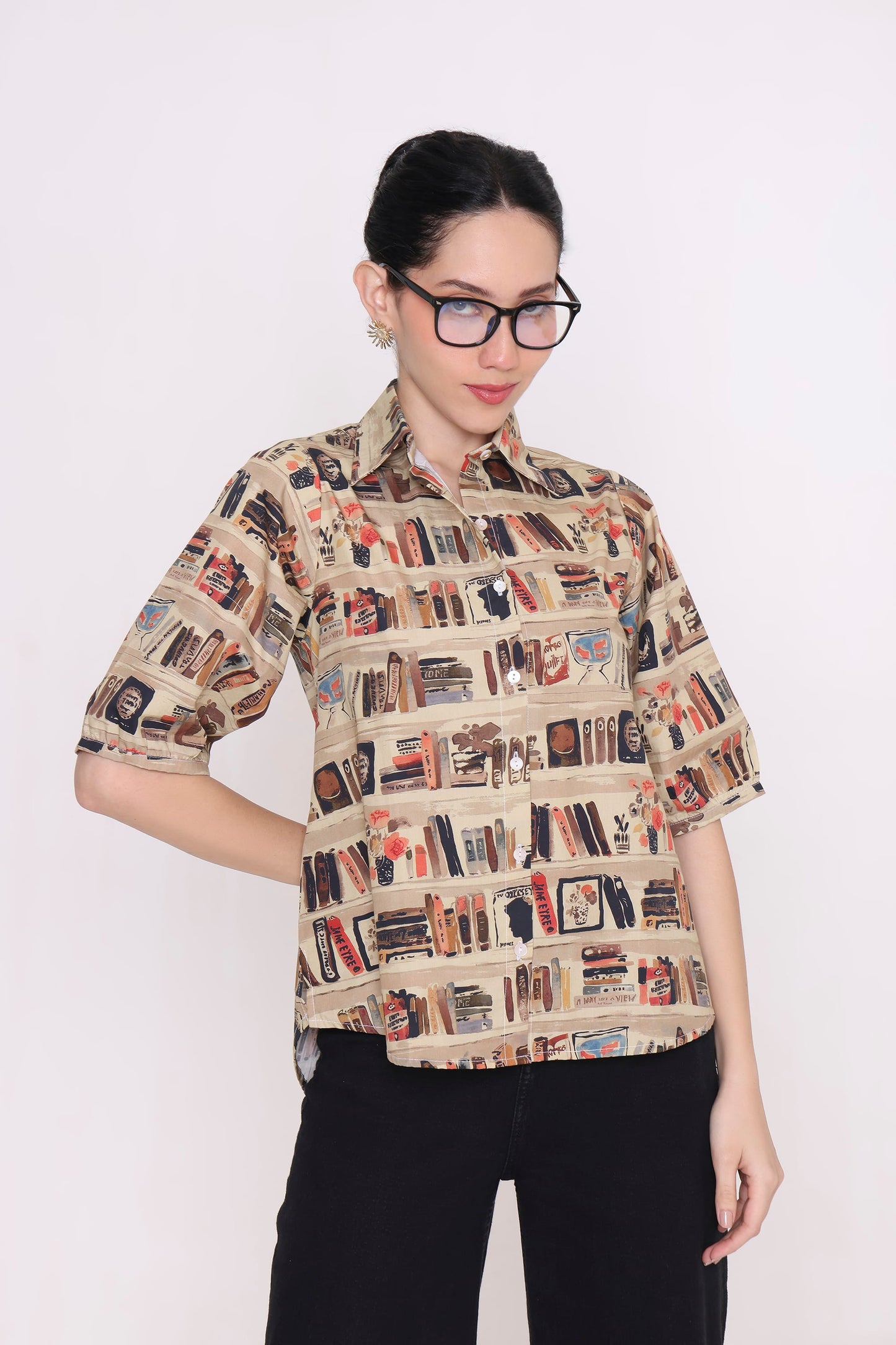 A woman wearing a beige short-sleeve button-up shirt with a detailed bookshelf and book pattern, paired with black pants, photographed against a plain white background.