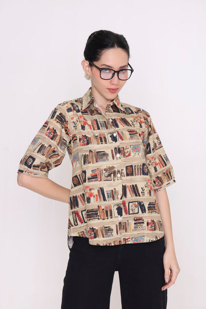A woman wearing a beige short-sleeve button-up shirt with a detailed bookshelf and book pattern, paired with black pants, photographed against a plain white background.