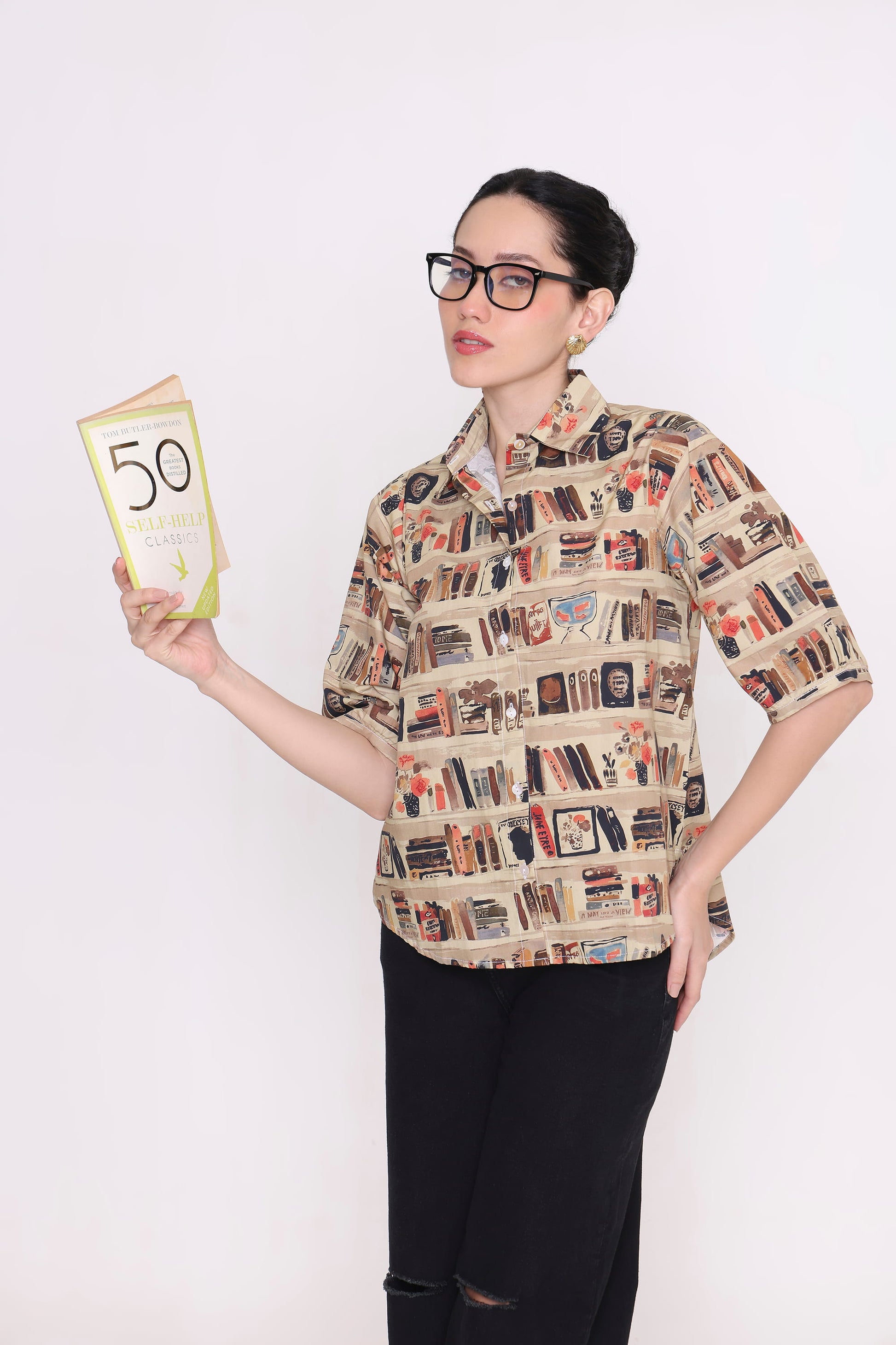 A woman wearing black-rimmed glasses and a beige short-sleeve button-up shirt with a detailed bookshelf pattern, holding a book titled '50 Great Quotes' against a plain white background.