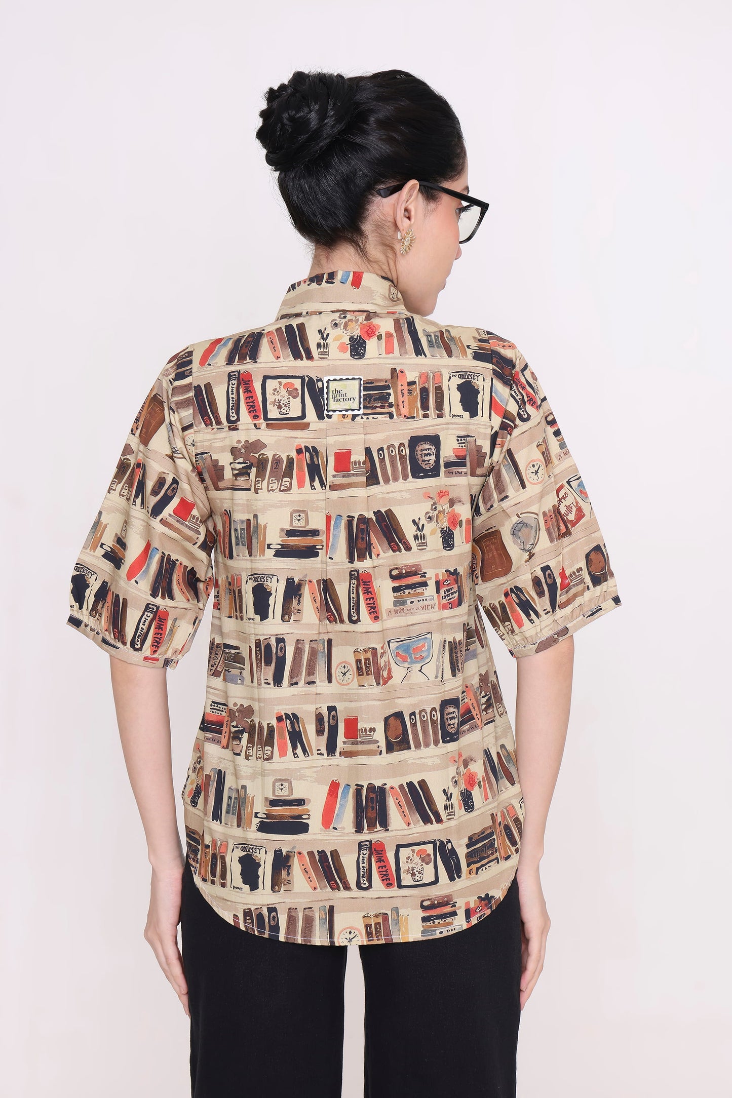 A woman viewed from the back wearing a short-sleeve button-up shirt with an all-over bookshelf and book print on a beige background, paired with black pants, against a plain white studio background.