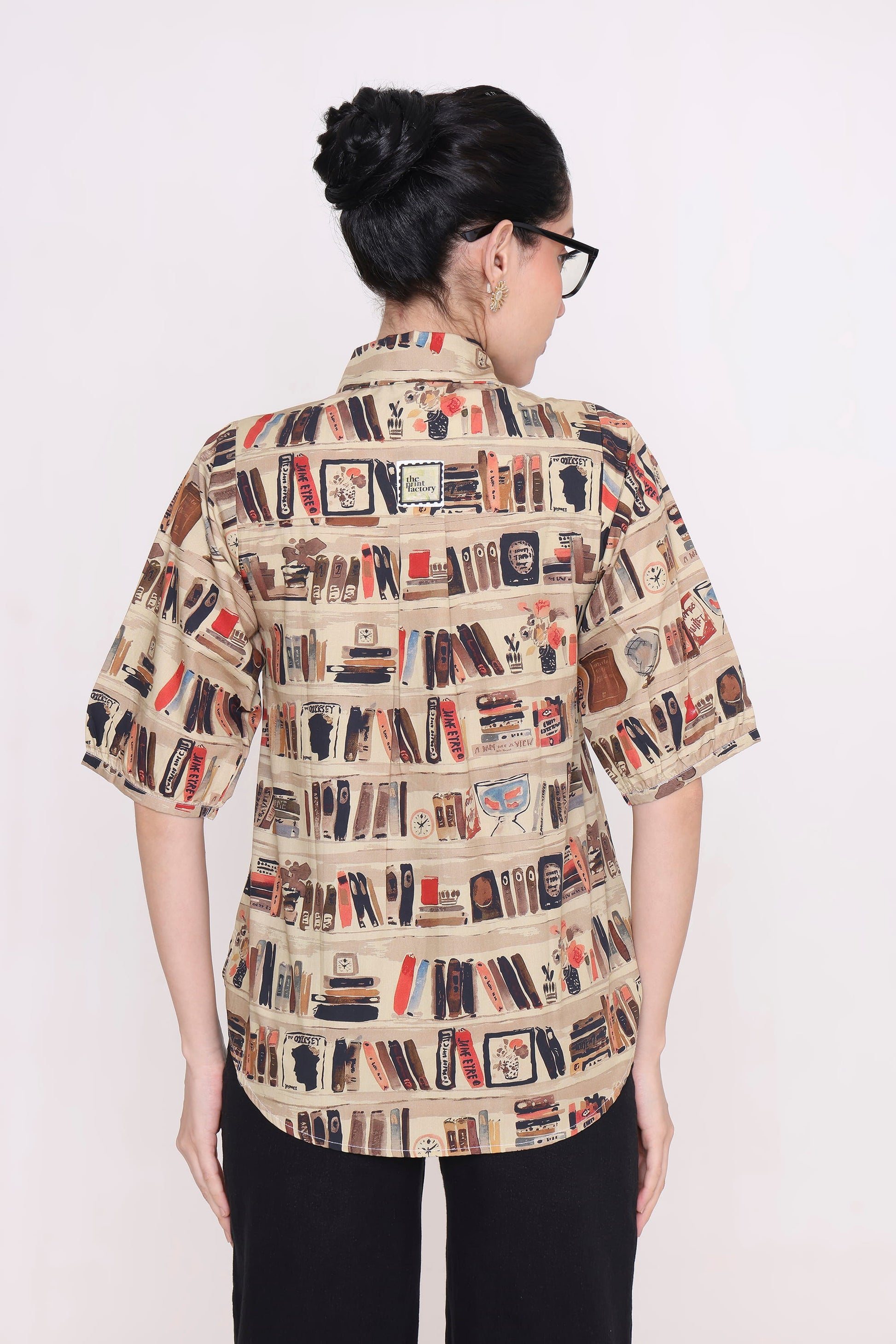 A woman viewed from the back wearing a short-sleeve button-up shirt with an all-over bookshelf and book print on a beige background, paired with black pants, against a plain white studio background.