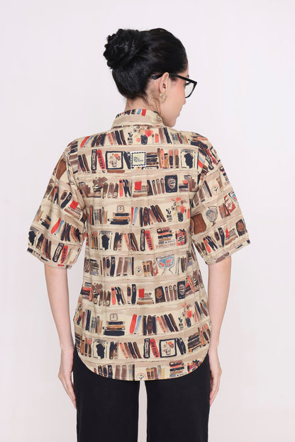 A woman viewed from the back wearing a short-sleeve button-up shirt with an all-over bookshelf and book print on a beige background, paired with black pants, against a plain white studio background.