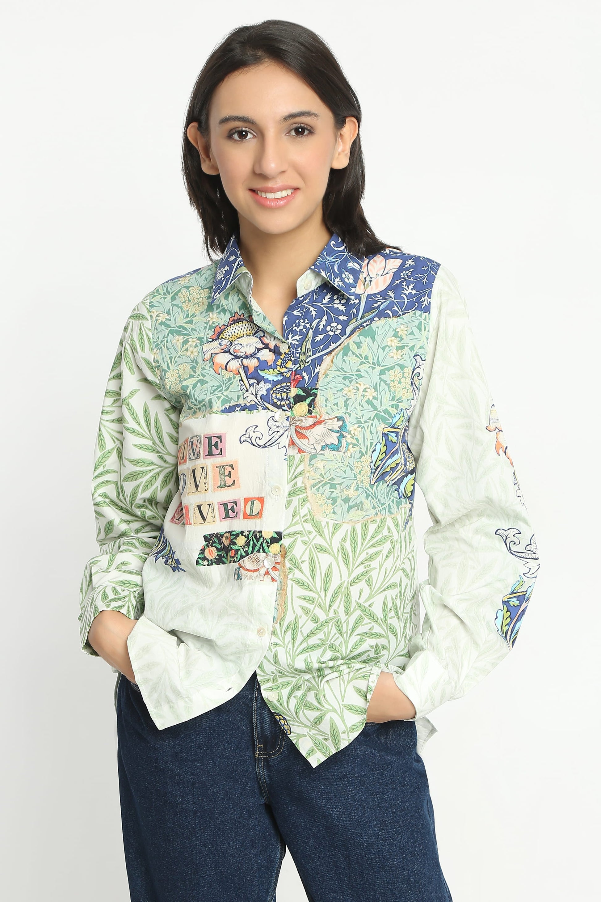 A woman modeling a long-sleeve button-up shirt with a multicolor patchwork print featuring green tropical leaves, blue floral details, and a white pocket with colorful text, paired with dark blue jeans against a plain white background.